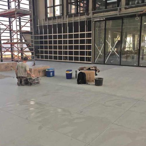Ein Handwerker bearbeitet den Betonboden in einem modernen Gebäude mit großen Glasfronten und Gerüstbau im Hintergrund.